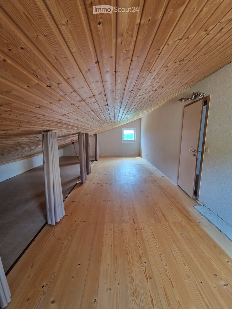 Empty room with wooden floor, wooden ceiling, and a small window at the far end. There is a door on the right side and a curtain on the left side.