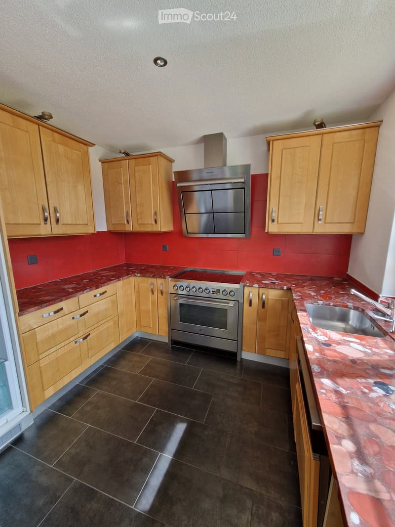 Modern kitchen with light wooden cabinets, stainless steel appliances, red tiled backsplash, and a marble countertop.
