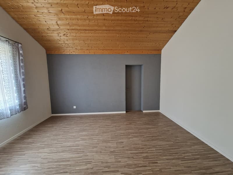 Empty room with wooden flooring, grey walls, and a wooden slanted ceiling. A single door is visible, and a curtain is partially drawn on the left side.
