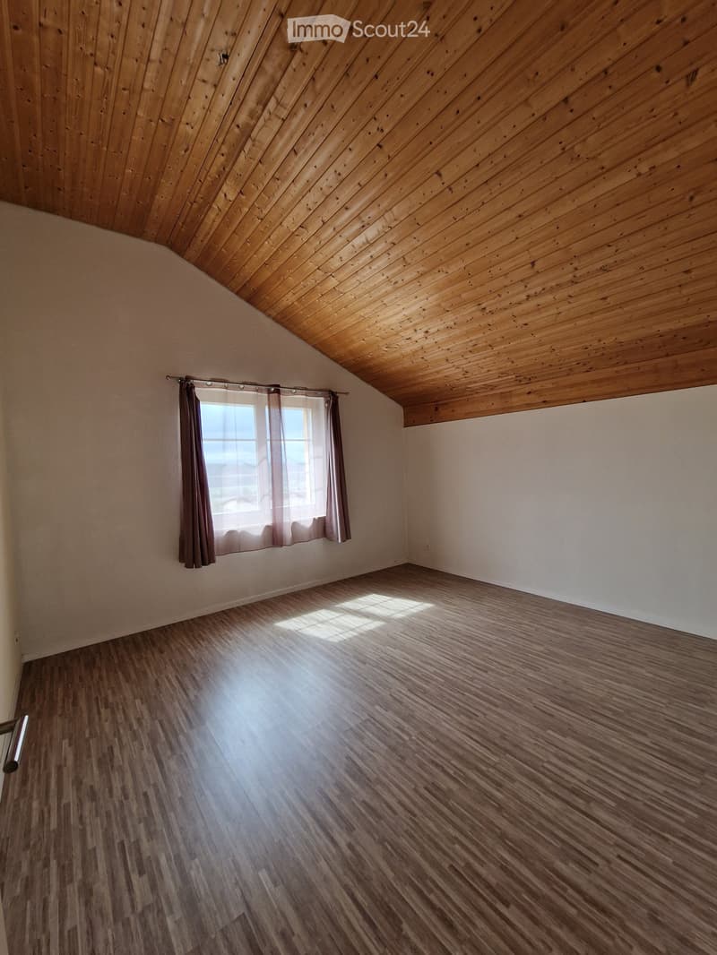 Empty room with wooden flooring, white walls, and a window with curtains. The ceiling is slanted and made of wood.