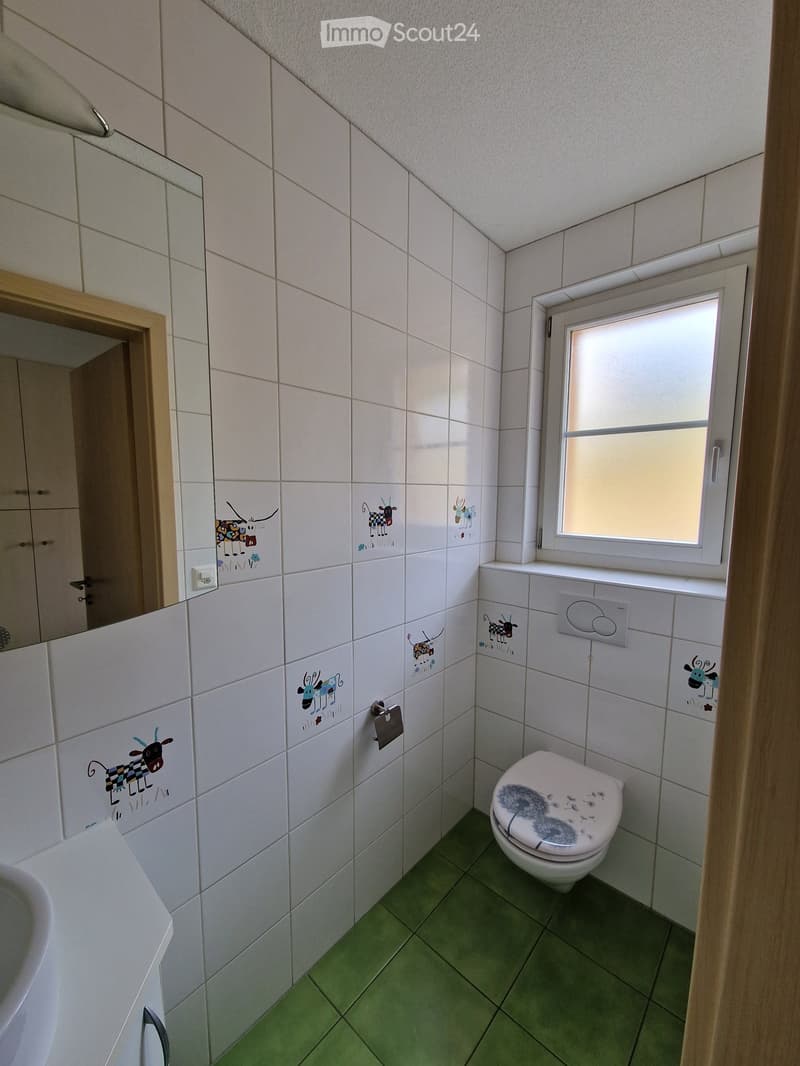 Small bathroom with white tiled walls, green floor tiles, a toilet with a decorative seat cover, a sink, and a mirror. The tiles feature cartoon animal decals.