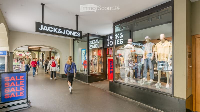Shopping mall corridor with people walking, storefronts including Jack & Jones, sale signs, and mannequins in display windows.