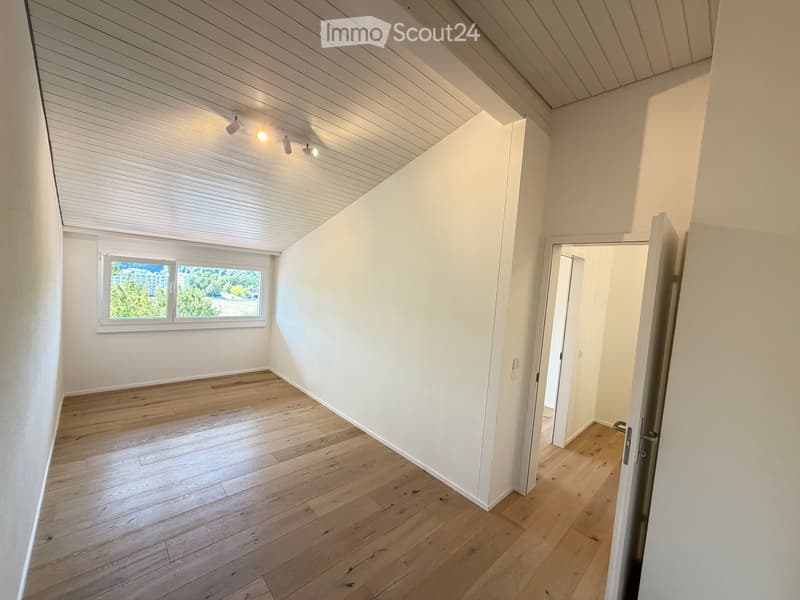 Empty room with wooden flooring, white walls, and a large window with blinds. The ceiling has recessed lighting and a slanted design, typical of an attic or loft space.