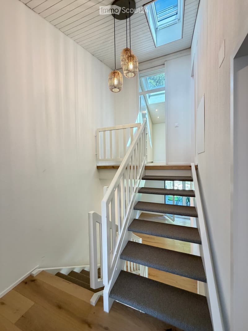 Modern staircase with wooden steps, white railing, and pendant lights, leading to a bright, open area with skylights.
