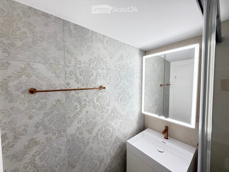 Modern bathroom with grey patterned tiles, a white sink with a copper faucet, and a large illuminated mirror.
