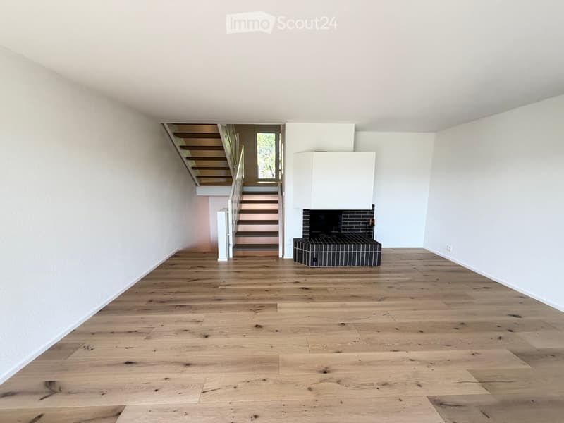Empty room with light wooden flooring, white walls, and a fireplace with a black tiled surround. A staircase leads to an upper level.