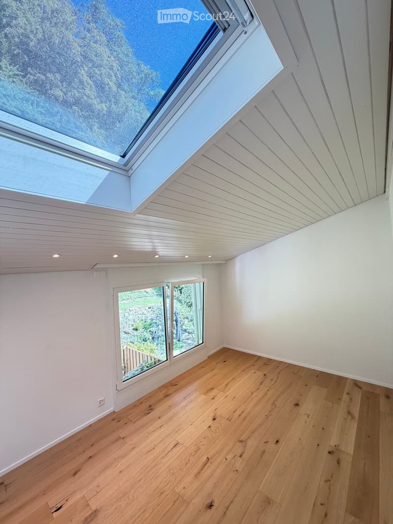 Empty room with light wooden flooring, white walls, and large windows including a skylight, offering natural light and a view of the outdoors.