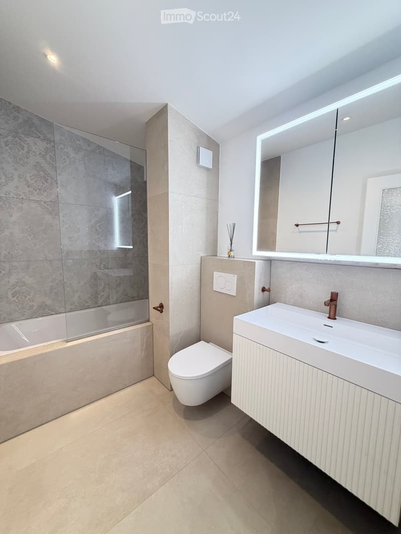Modern bathroom with a bathtub, toilet, sink, and large mirror. Features light-colored tiles and a sleek design.