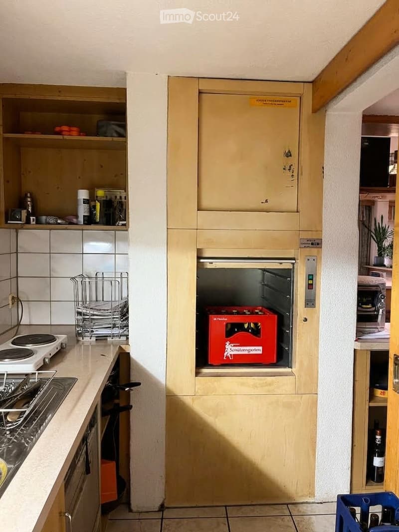 The kitchen features an induction stove, a dish rack, and an open oven with a red box inside. Shelves hold various items, including bottles and a camera, and there is a bottle on the floor near the oven.