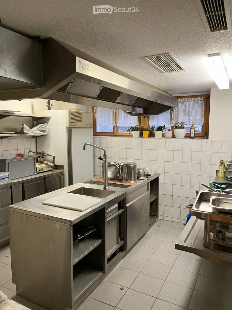 Commercial kitchen with stainless steel counters, sink, cutting board, refrigerator, microwave, oven, and various pots and pans. The space features tiled walls and floors, a large window with potted plants, and a metal vent hood above the cooking area.