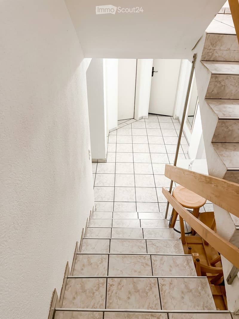 A narrow hallway with tiled flooring and white walls, featuring a staircase leading upwards and a wooden chair placed against the wall.