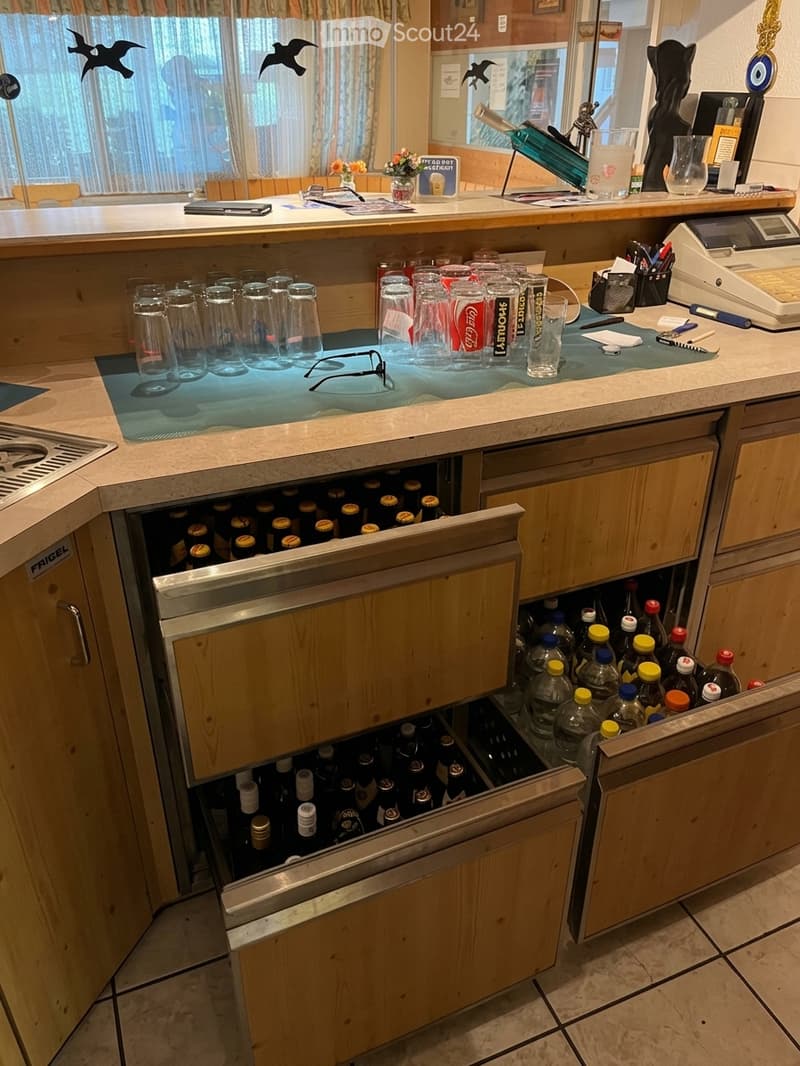 A modern kitchen with a wooden countertop, multiple drawers filled with bottled drinks, and a variety of glasses neatly arranged. A cash register, pen, notepad, and a vase with flowers are also visible on the counter.