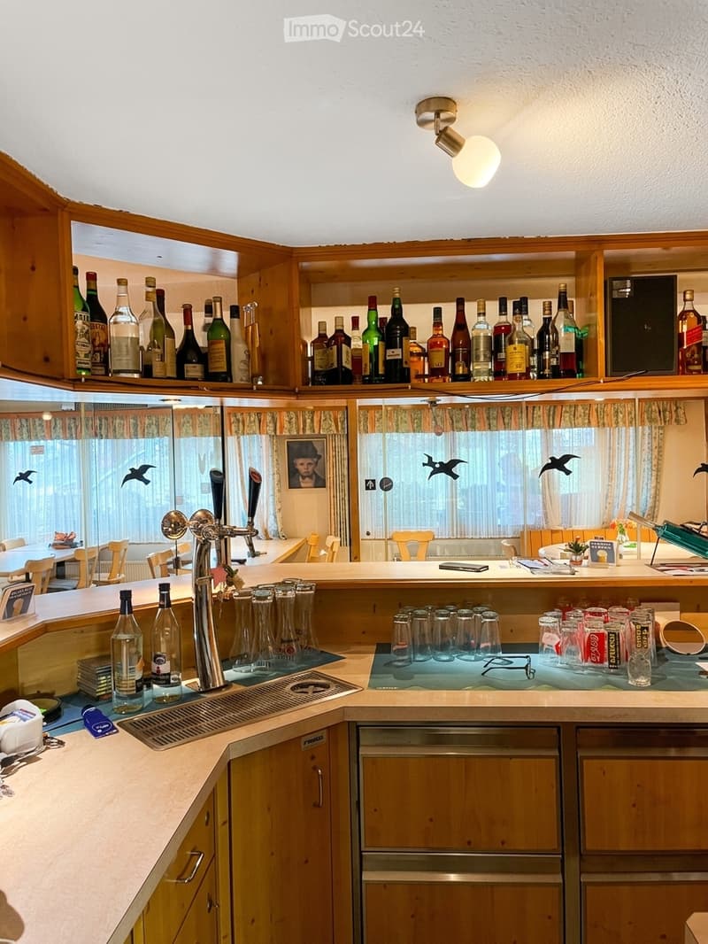 A cozy kitchen with wooden cabinets, a sink, a faucet, and shelves stocked with various bottles. The counter features glasses, a faucet, and a few decorative items, with a dining area visible through a large window.