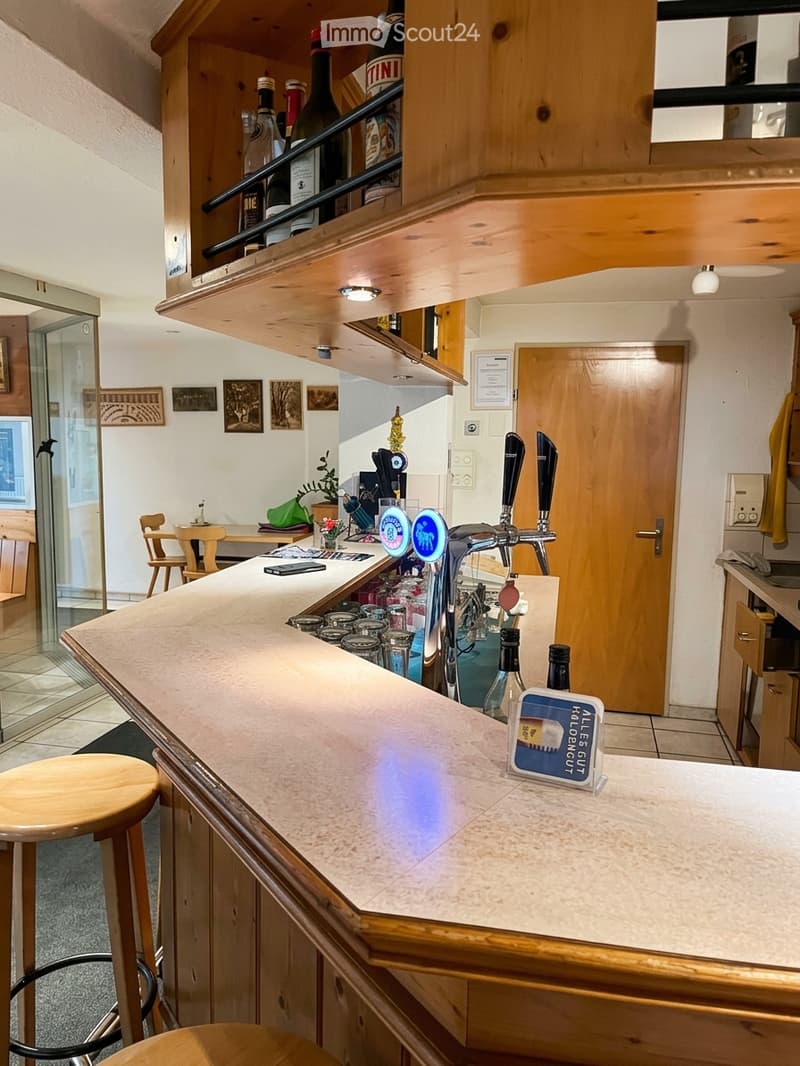 A modern bar area with a polished wooden counter, beer taps, bar stools, and shelves stocked with bottles. There is a small table with chairs, a green bag, and various bar accessories.