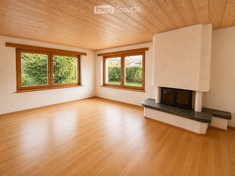 Empty room with wooden flooring, white walls, and a fireplace with a dark stone surround. Two large windows with wooden frames offer views of greenery outside.