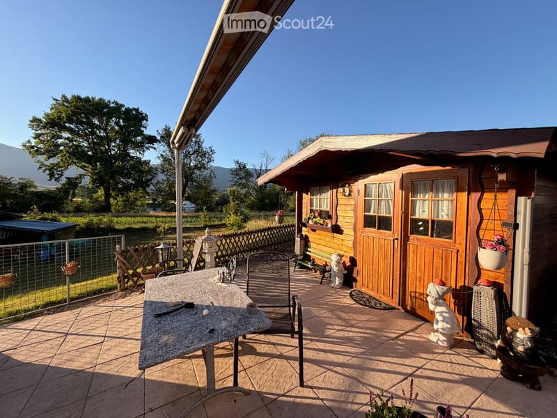 A wooden cabin with a covered patio area, featuring a stone-tiled floor, a table with chairs, garden ornaments, and a view of a green landscape with trees and mountains.