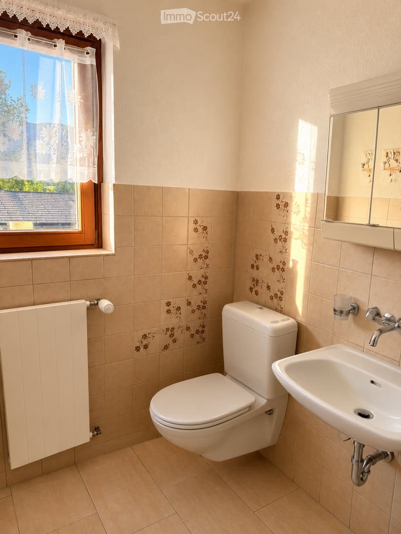 Bathroom with beige tiles, a toilet, a sink, a mirror, and a radiator. The window has white lace curtains.
