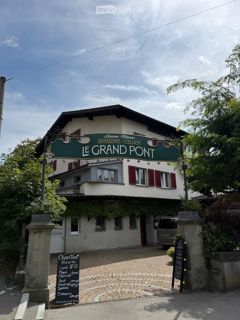 Two-story building with white walls, red shutters, and a green sign reading "Le Grand Pont." A van is parked in front, and there are stone pillars and a paved entrance area.