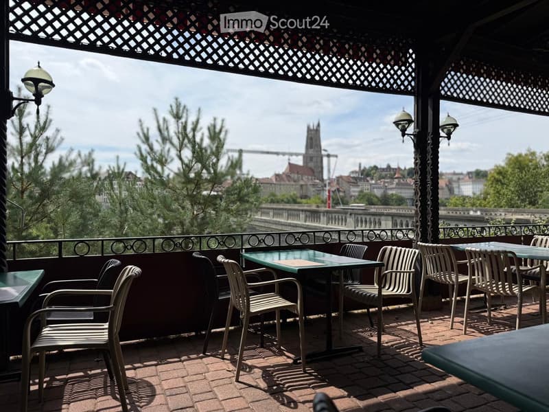 An outdoor terrace with tables and chairs, overlooking a cityscape with trees and a distant church tower.