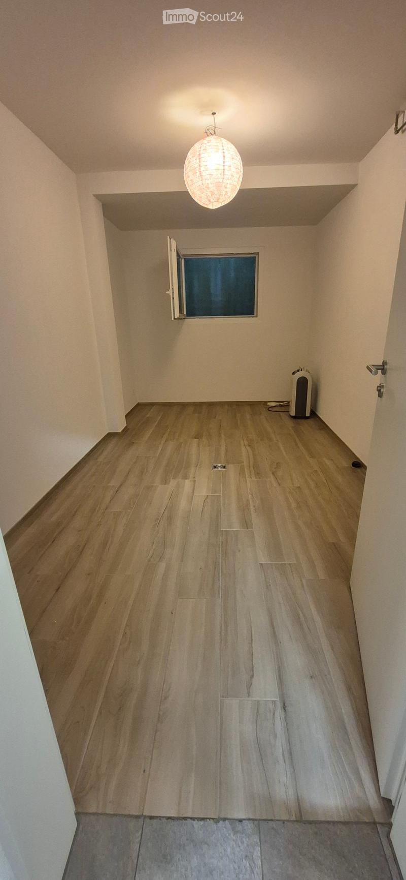 Empty hallway with wooden flooring, white walls, a hanging light fixture, and a small window with an open door.