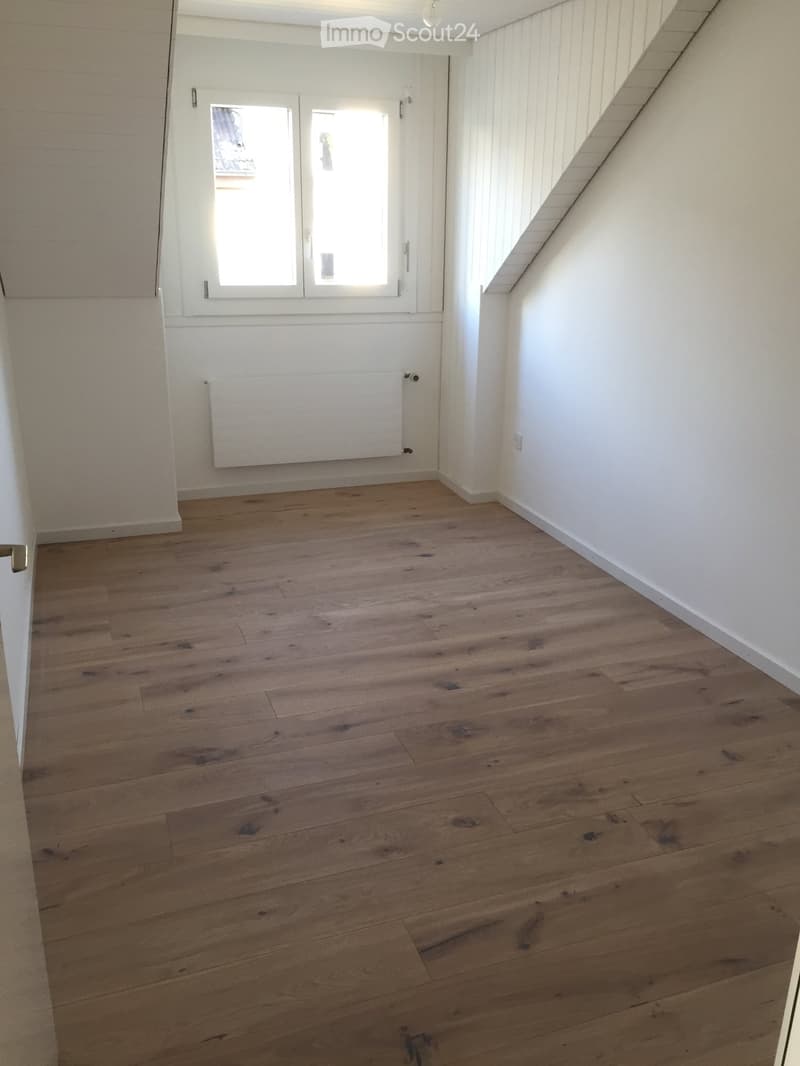 Empty room with wooden flooring, white walls, and a window with white frames. A radiator is mounted on one wall.