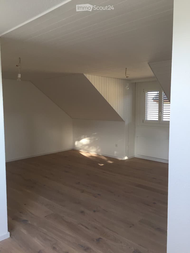 Empty room with wooden flooring, white walls, and a slanted ceiling. Features a window with blinds and exposed wooden beams on the ceiling.