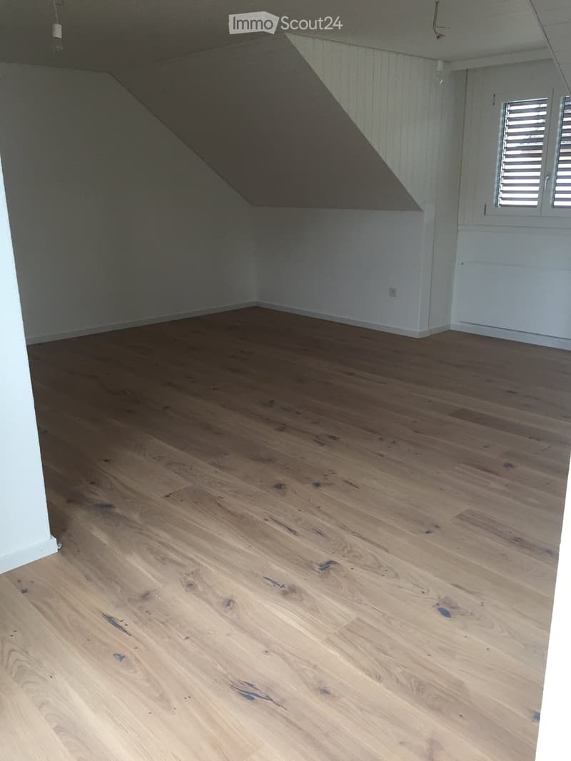 Empty room with wooden flooring and white walls, featuring a slanted ceiling and a window with shutters.