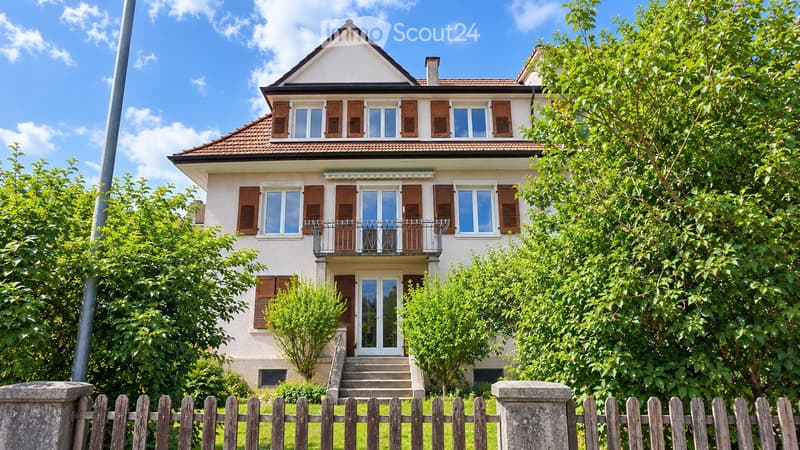 Two-story house with beige walls, brown shutters, and a tiled roof. Features a balcony on the upper floor and a small garden with a wooden fence.