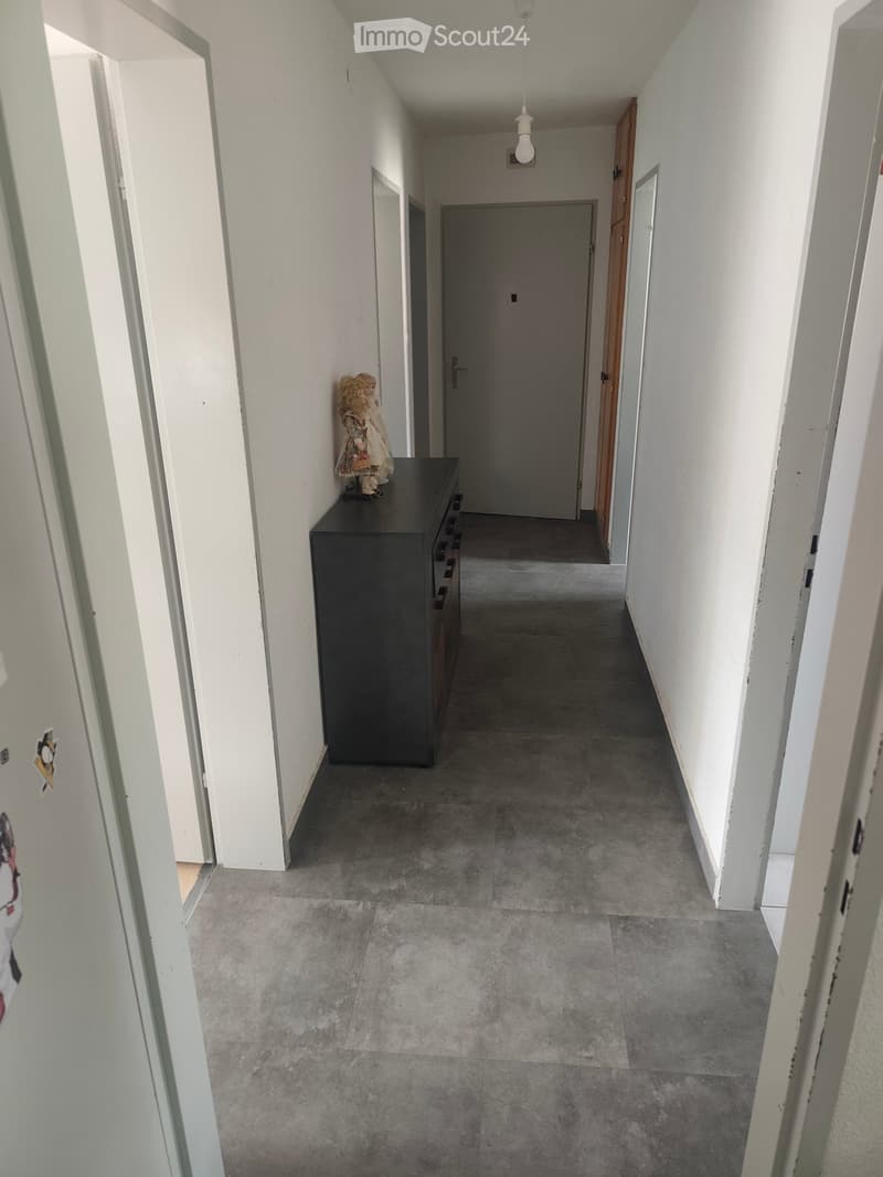 Hallway with grey tiled floor, white walls, a black cabinet with drawers, and a decorative doll on top.