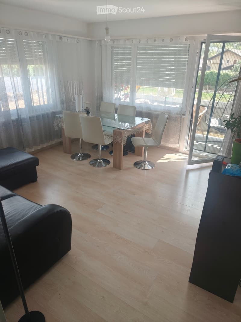 Bright living and dining area with light wooden flooring, large windows with blinds, a black leather sofa, a glass dining table with four white chairs, and a small decorative table runner.