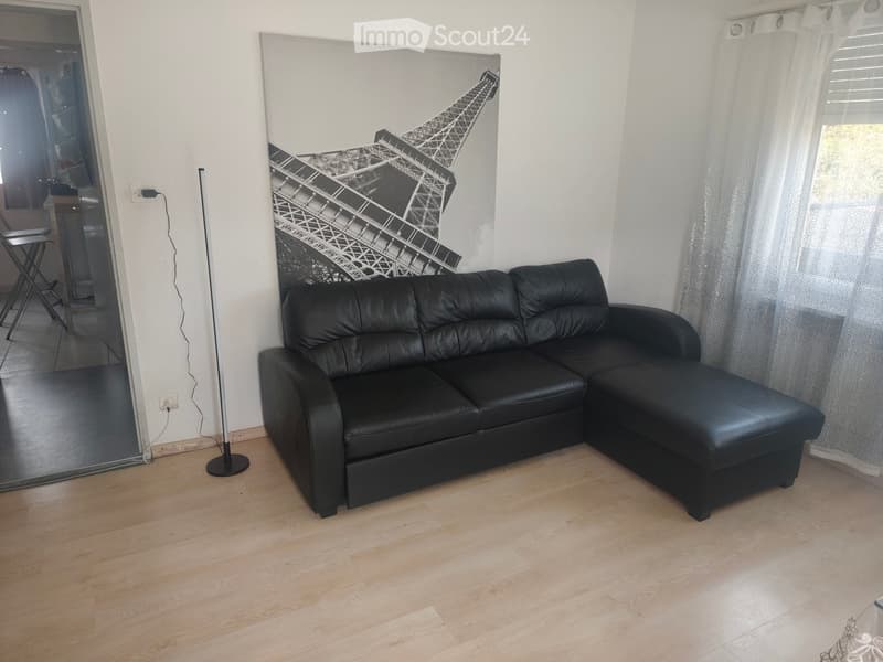 Modern living room with light wooden flooring, a black leather sectional sofa, a floor lamp, and a large Eiffel Tower mural on the wall. A window with white curtains is visible on the right side.