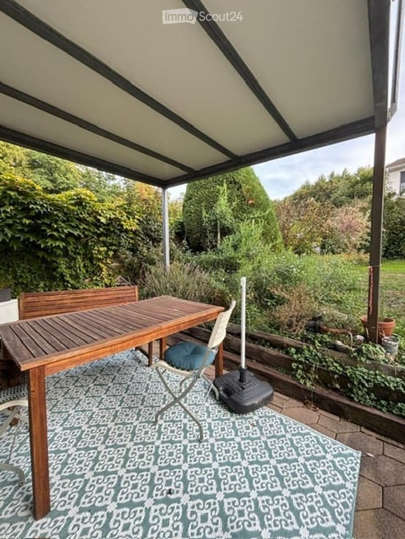 A covered outdoor patio with a wooden table, chairs, and a patterned rug, surrounded by greenery and a garden area.