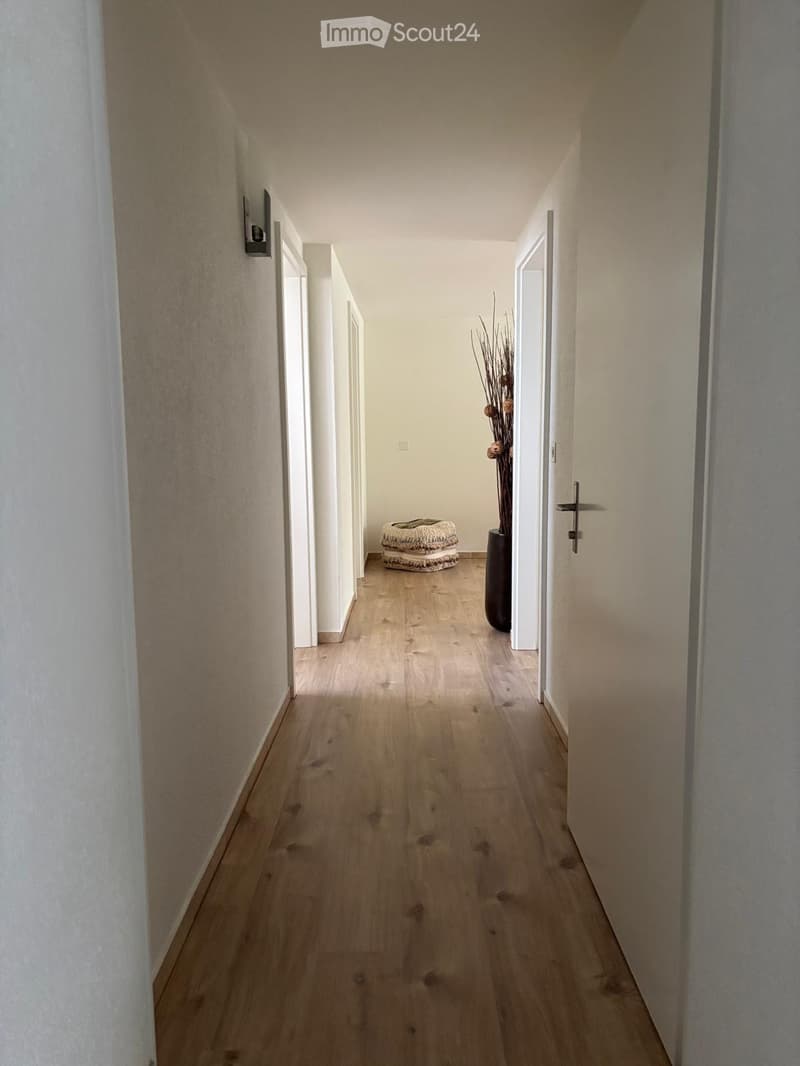 A long, narrow hallway with light wooden flooring, white walls, and several doors leading to other rooms. A tall vase with dried plants is placed near the end of the hallway.