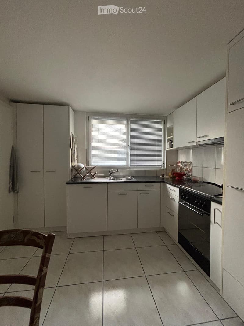 Modern kitchen with white cabinets, tiled floor, and stainless steel appliances including a sink, faucet, and oven. A dish rack and a wooden chair are visible.
