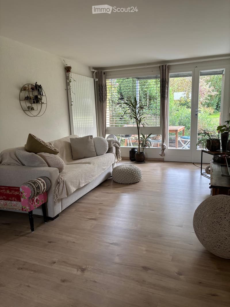 A modern living room with a large sofa, ottoman, and a small table near the window. The room features wooden flooring and large windows with blinds, leading to a balcony area with outdoor furniture.