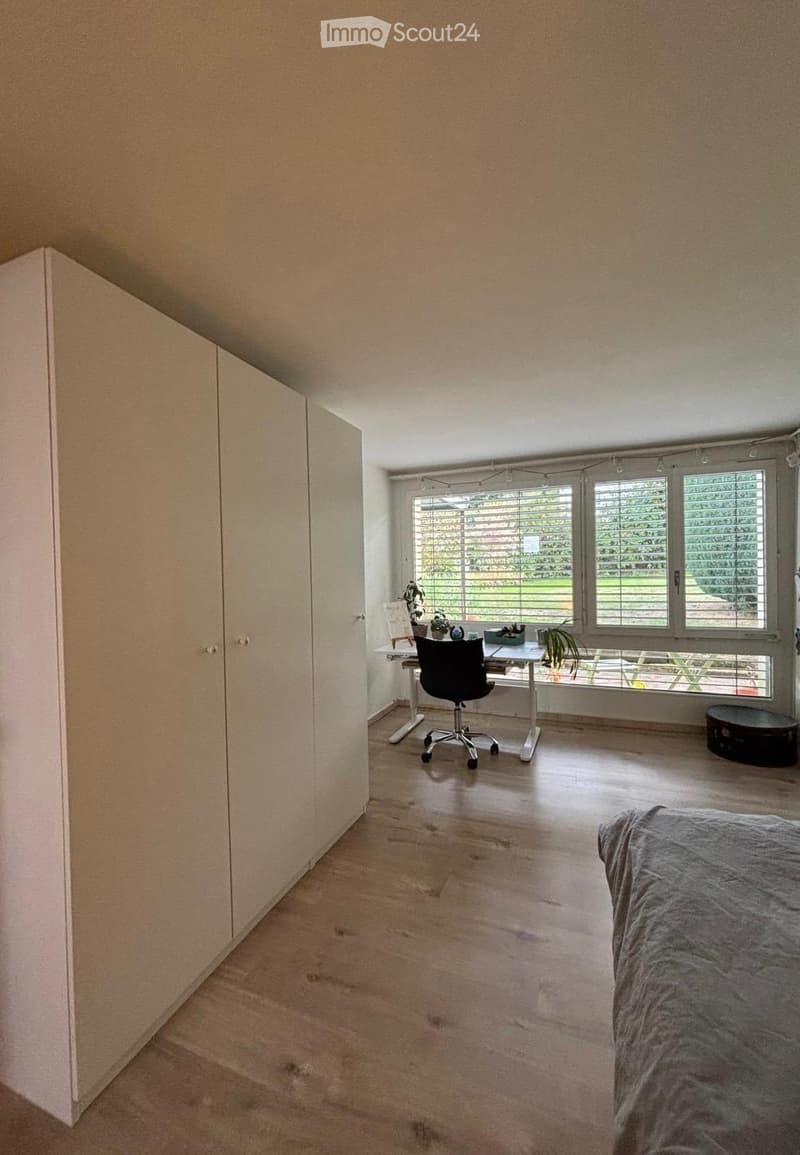 Bright bedroom with a large bed, a tall wardrobe, a desk with a chair, and a small stool near the window. The room has wooden flooring and large windows with blinds.