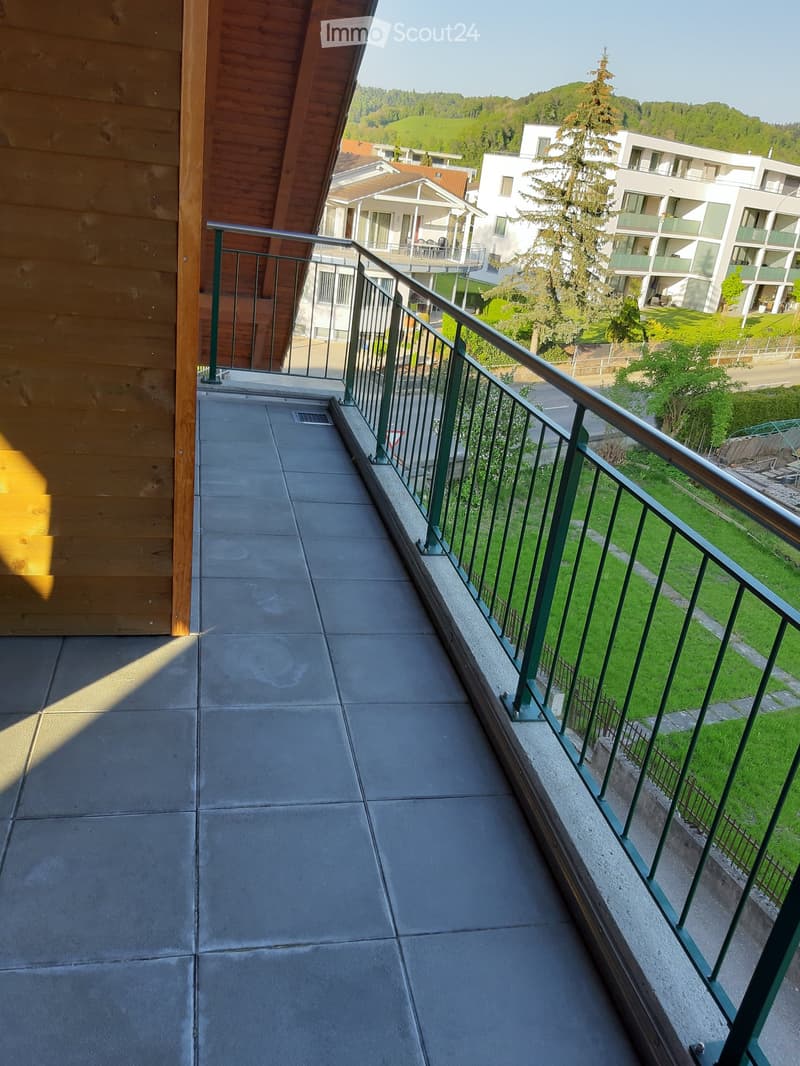 Balcony with green metal railing, concrete floor, and view of a residential area with buildings and greenery.