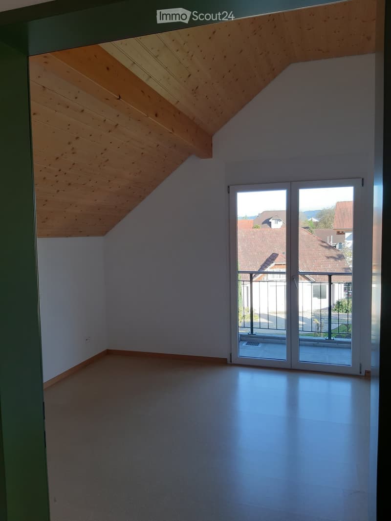 Empty room with wooden slanted ceiling, white walls, and a large sliding glass door leading to a balcony with a metal railing.