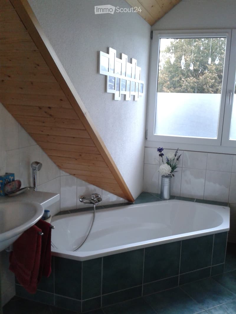 Bathroom with a bathtub, sink, and a vase with flowers on the windowsill. The room features wooden slanted ceilings and a large window with a view of trees.