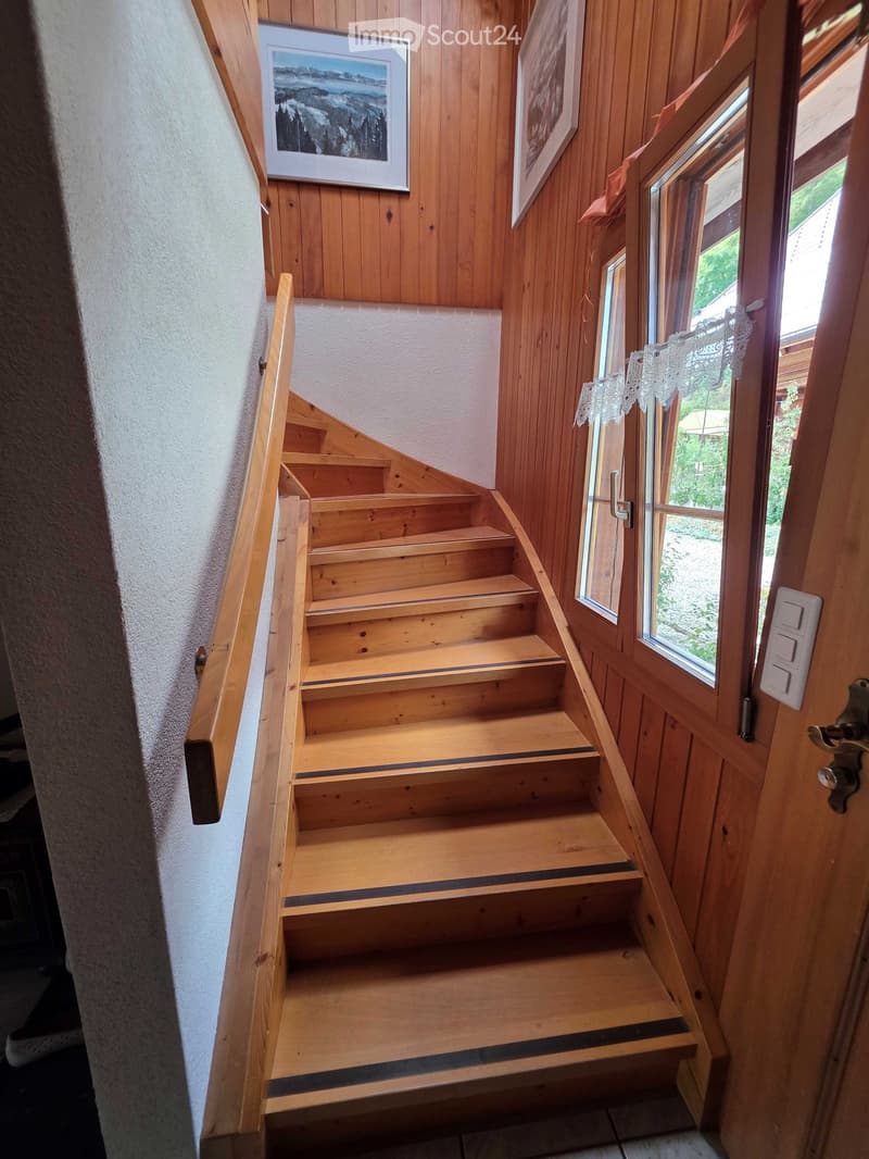 Wooden staircase with handrail, white wall, framed pictures, and a window with lace curtains.