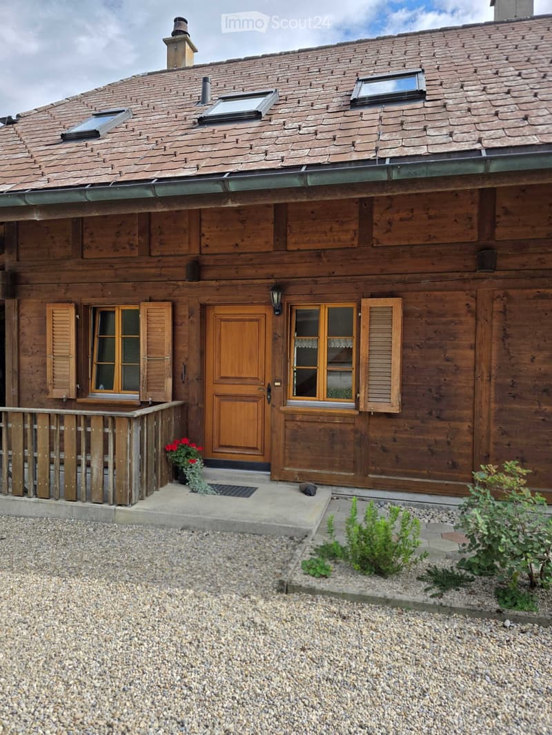 A two-story wooden house with a sloped roof, multiple windows, and a wooden front door. The house features a small porch with a wooden railing and a potted plant near the entrance.