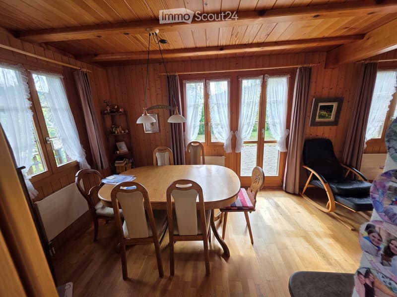 Cozy wooden-paneled dining room with a round wooden dining table surrounded by six chairs, a black leather sofa, and a small shelf with decorative items.