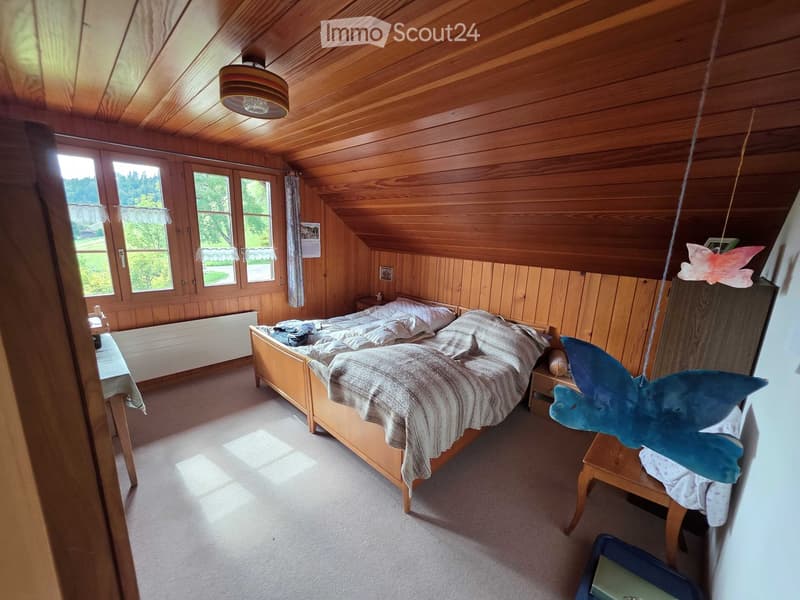 Cozy attic bedroom with wooden walls and ceiling, large windows, a double bed with pillows and blankets, nightstands, and decorative items.