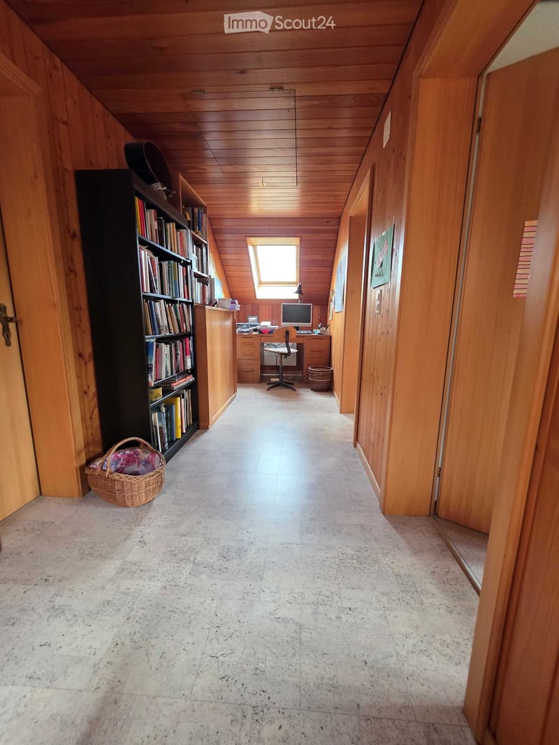 Wooden-paneled hallway with a small office area visible at the end, featuring a desk, chair, and bookshelf. Natural light from a skylight illuminates the space.