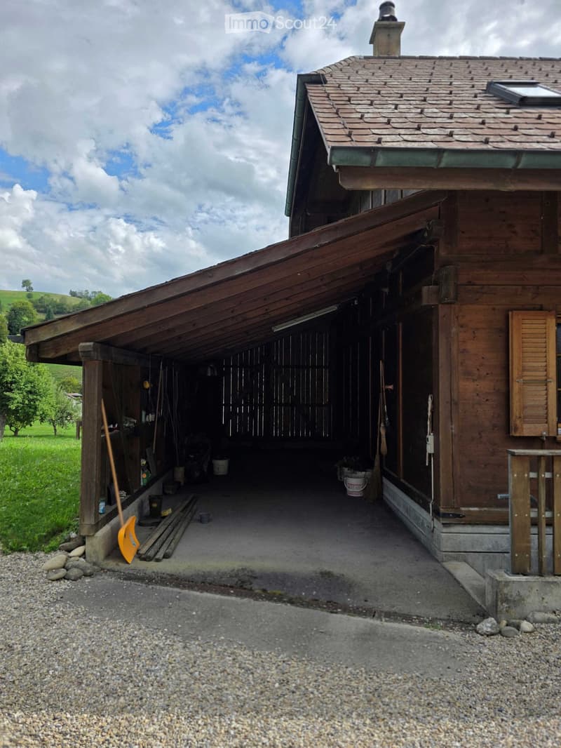Wooden barn with a large open garage area, tools, and storage items visible inside. The building has a sloped roof and is situated in a rural, green landscape.