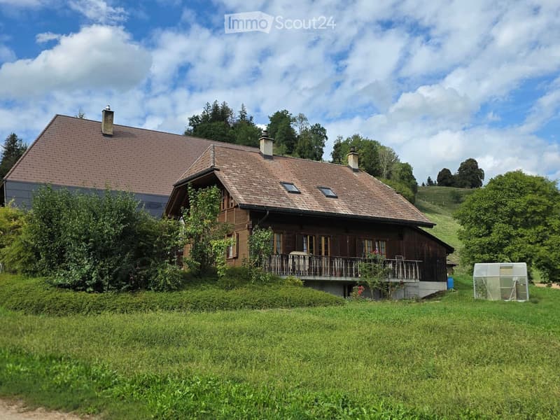 A two-story wooden house with a brown tiled roof, multiple chimneys, and a balcony overlooking a grassy lawn. The house is surrounded by trees and greenery.