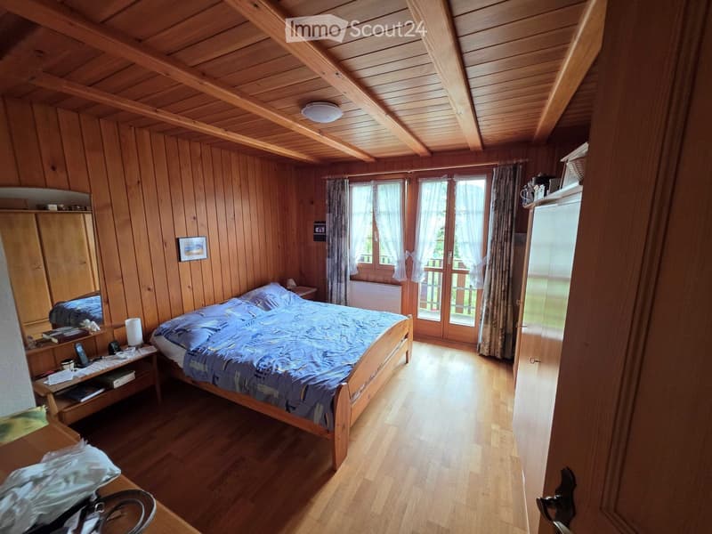 Cozy wooden bedroom with slanted wooden ceiling, large bed with blue bedding, nightstand, mirror, and wooden furniture.