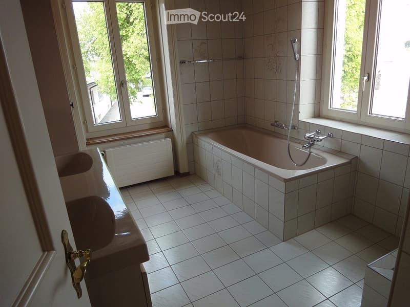 Bathroom with a bathtub, sink, and radiator. The room has tiled walls and floor, with windows providing natural light.