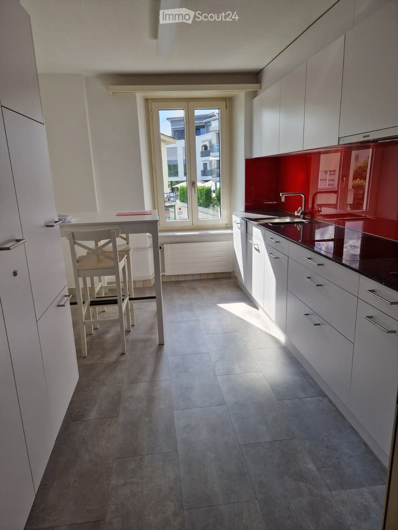 Modern kitchen with white cabinets, red backsplash, and a dining area with a table and chairs. Large windows offer a view of the outside.