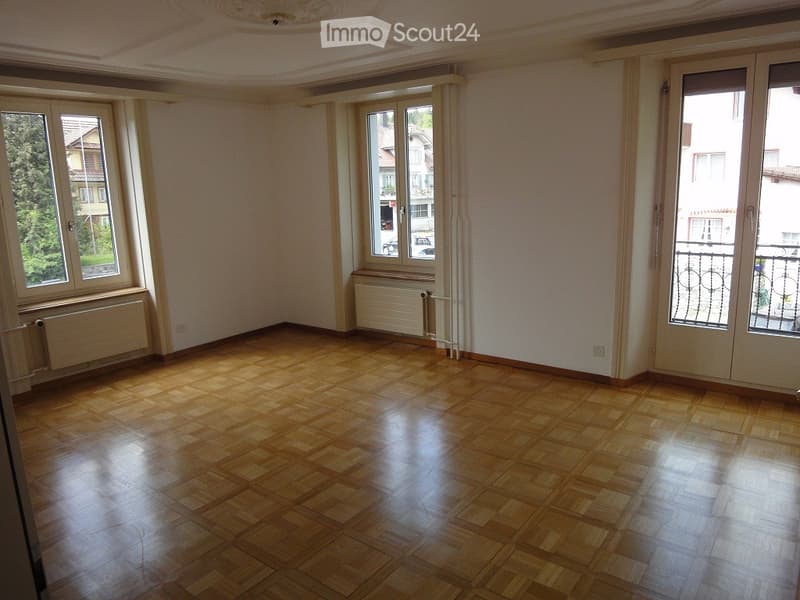 Empty room with wooden flooring, large windows, radiators, and a balcony door leading to a small balcony area.